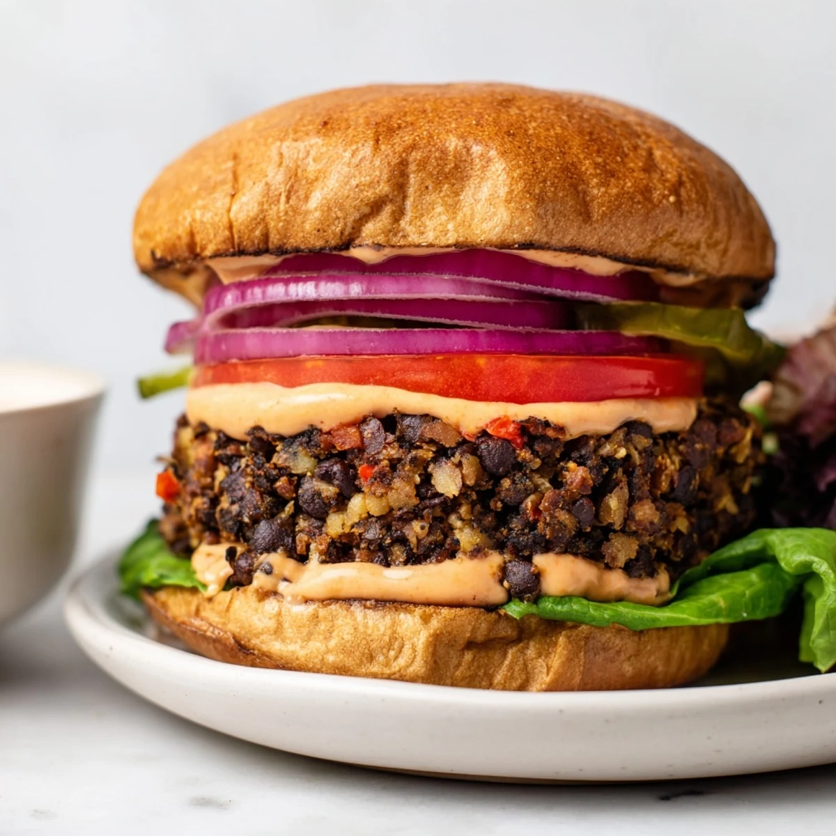 Spicy chipotle mayo is drizzled over a Vegan Black Bean Burger, paired with golden sweet potato fries on a rustic wooden platter.