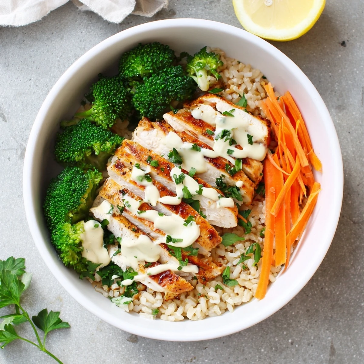Sliced grilled chicken breasts arranged over steamed broccoli and brown rice drizzled with white creamy garlic sauce and fresh parsley